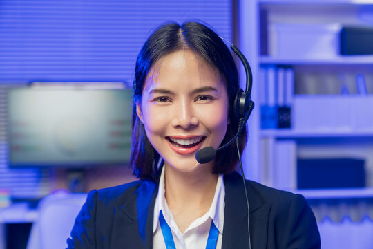 Smiling young Asian customer service representative wearing microphone headset and working on laptop in modern office.
