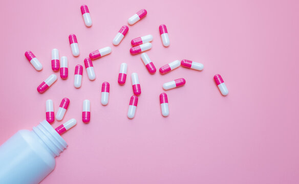 Pink-white capsules pill spilled out from plastic bottle container on pink background. Antibiotics drug resistance. Antimicrobial capsule pills. Pharmaceutical industry. Pill for happy love concept.