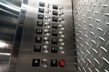 A close-up photograph of a metallic industrial elevator control panel featuring rows of numbered buttons with integrated braille, a red emergency stop button, and an alarm signal, set against a rugged © kareem