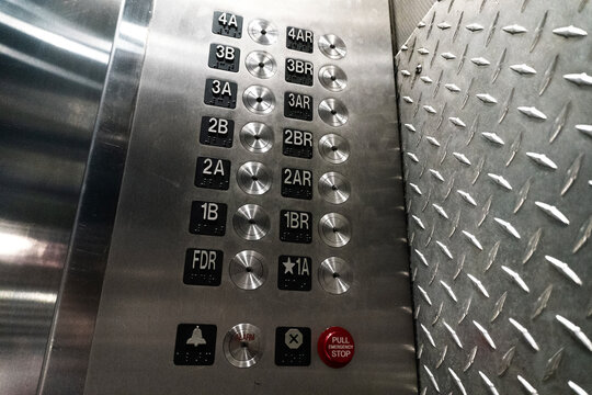 A close-up photograph of a metallic industrial elevator control panel featuring rows of numbered buttons with integrated braille, a red emergency stop button, and an alarm signal, set against a rugged