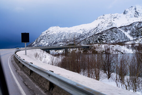 POV of driving a car on an empty winding wet asphalt highway stretching through a vast snow-covered Arctic landscape towards distant mountains under a cloudy sky in winter Scandinavia.