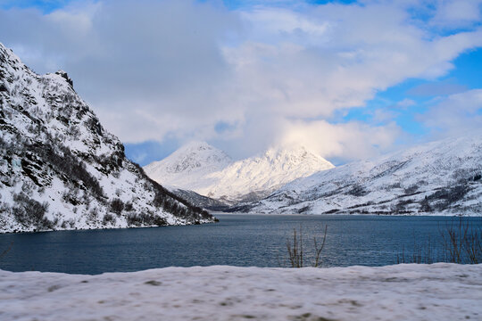 Scenic POV of driving a car approaching a concrete bridge across a winter fjord in Lofoten, Norway. Beautiful landscape with majestic snow-covered mountains under a dramatic sky on a Nordic road trip.