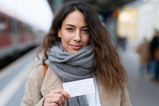 Woman smiles gently, holding train ticket. Wearing cozy scarf and soft coat. Platform behind her, train passing by. Background blurred, emphasizing her calm expression