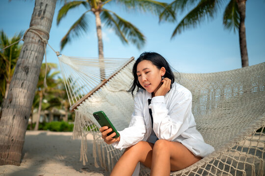 Happy young woman talking on her smartphone at a tropical beach, enjoying vacation, remote work