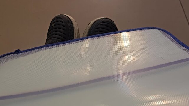 Close-up of feet in dark slip-on shoes resting behind a clear plastic zipper folder with blue trim on a tiled floor