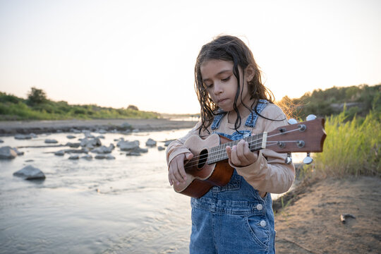 Young girl playing ukulele by a river at sunset