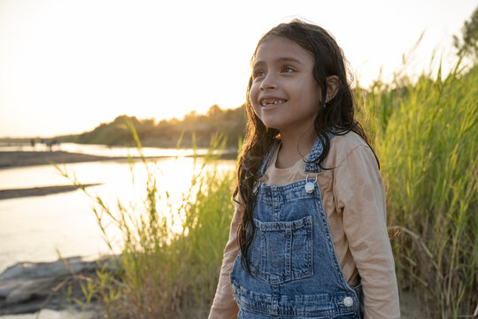 Young girl in overalls smiles by a river at sunset