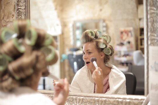 Woman with hair rollers applying makeup in mirror
