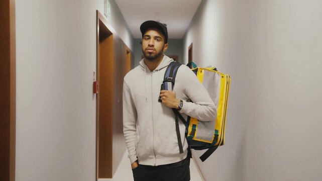 Young delivery man standing in apartment hallway wearing yellow food delivery backpack