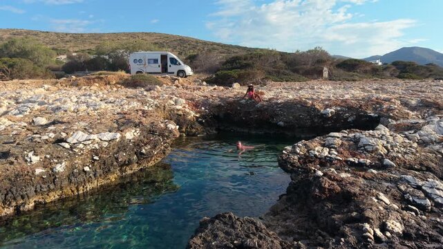 Explore the breathtaking rocky coastline of Paros Island, Greece with shimmering turquoise waters. A couple relaxes by the ocean natural pool, a man and a woman from above drone view with a camper van