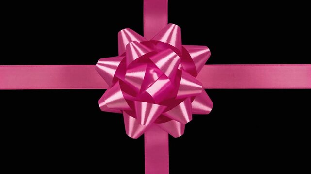 A shiny pink ribbon bow at the intersection of two pink ribbon strips arranged in a cross pattern against a black background, reflecting light to emphasize loops, folds, and celebratory symbolism.