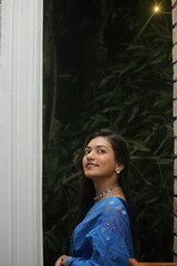 Fototapeta premium Graceful woman in blue sari and jewelry posing next to a white pillar against dark foliage background