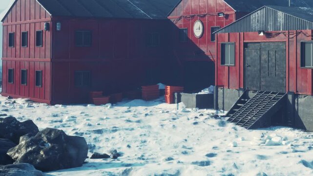 Red structures stand on a snowy landscape near the water. The ice covers the ground, and the buildings appear to be part of a research station. Snow surrounds the area.