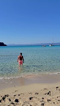 A man walks through the soft sand towards the mesmerizing turquoise waters of Simos Beach. Sunlight sparkles on the waves, inviting relaxation and adventure in this stunning Elafonisos Island, Greece