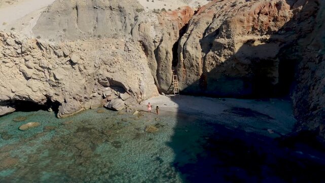 A couple of men and woman walks at the Tsigrado Beach in Milos, Greece. The soft white rocks contrast beautifully with the crystal-clear waters, inviting visitors for a perfect escape into nature