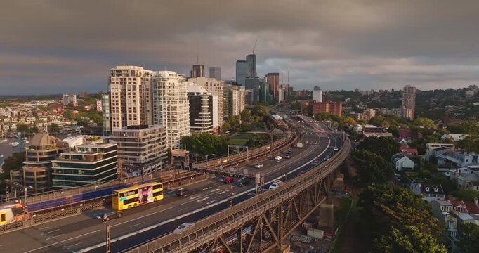 Sydney City skyline and North Sydney connecting with the Harbour Bridge, showing vehicles cars driving on the highway and a train moving alongside urban buildings in the morning light. Static shot