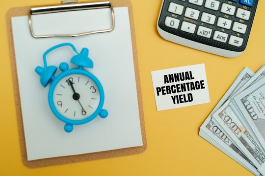 A blue alarm clock on a clipboard with "ANNUAL PERCENTAGE YIELD" text, calculator and US dollar notes on a yellow background. Concept of savings, interest growth and financial planning