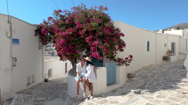 A diverse couple explores the picturesque streets of Lefkes, Paros, Greece, where a traveler enjoys a moment beside a beautiful bougainvillea. Enjoy the warm sunshine and tranquil atmosphere 