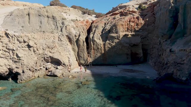 A couple of men and women walk at the Tsigrado Beach, Milos, Greece, where sun-drenched cliffs meet tranquil turquoise waters. Perfect for relaxation, swimming, and soaking in natures beauty