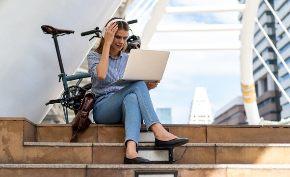 Urban Commute: A young woman wearing headphones works on her laptop while sitting on outdoor steps next to her foldable bicycle, blending technology and active urban lifestyle.