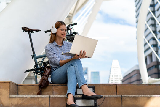 Urban Commute: A young woman wearing headphones works on her laptop while sitting on outdoor steps next to her foldable bicycle, blending technology and active urban lifestyle.