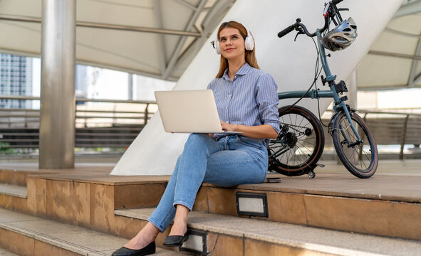 Urban Nomad: A young woman with headphones on, engrossed in her laptop, takes a moment to relax outdoors next to her foldable bicycle, embracing a modern, on-the-go lifestyle amid city architecture.