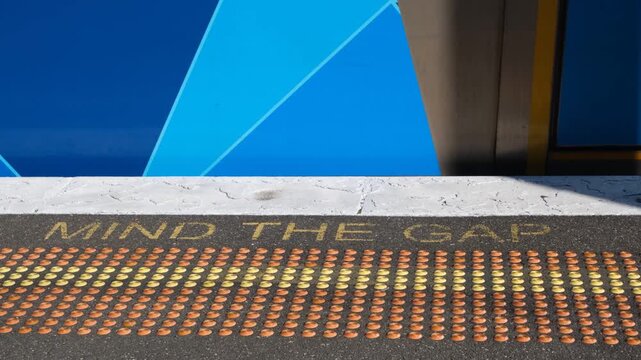 "Mind the Gap" safety text is painted on a train platform edge beside tactile paving against a moving train. Public transport safety at a railway station