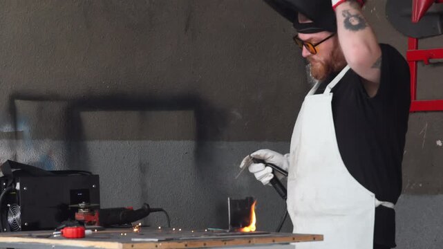 Close up of a welder in mask performing arc welding with sparks and blue smoke in workshop