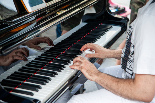 Una persona suona un pianoforte all'aperto. Il primo piano si concentra sulle sue mani sui tasti bianchi e neri, che si riflettono sulla superficie lucida del pianoforte.