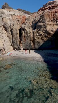 A couple discover the enchanting beauty of a secluded Tsigrado Beach in Milos, Greece. Towering cliffs frame the turquoise waters, where visitors enjoy the sun of this idyllic paradise
