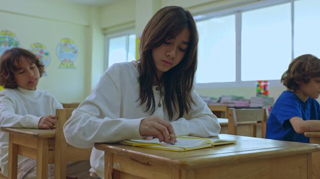 Asian girl classroom student desk notebook writing child school education pencil focused learning study young student at wooden desk among classmates in bright elementary room beside window