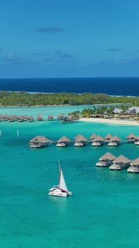 Vertical Screen: Bora Bora tropical paradise with white catamaran sailing through turquoise lagoon waters past rows of overwater bungalows and palm-covered islands under clear blue sky.
