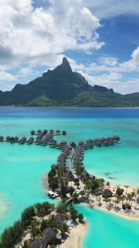 Vertical Screen: Bora Bora tropical paradise featuring overwater bungalows in turquoise lagoon with Mount Otemanu volcanic peak rising against blue sky with white clouds.