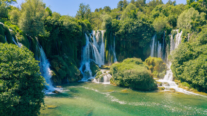 Beautiful cascading waterfall flowing into crystal clear emerald river surrounded by lush forest © Sanja
