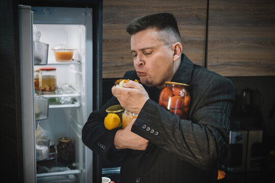 Overweight man in a suit eating a sandwich in front of an open fridge, holding jars of food. Gluttony, unhealthy eating, and compulsive snacking concept.
