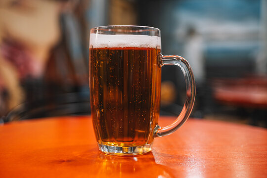 A glass mug of amber beer with a white foam head on a polished wooden table. Bar atmosphere, cold refreshment, and classic pub drink.