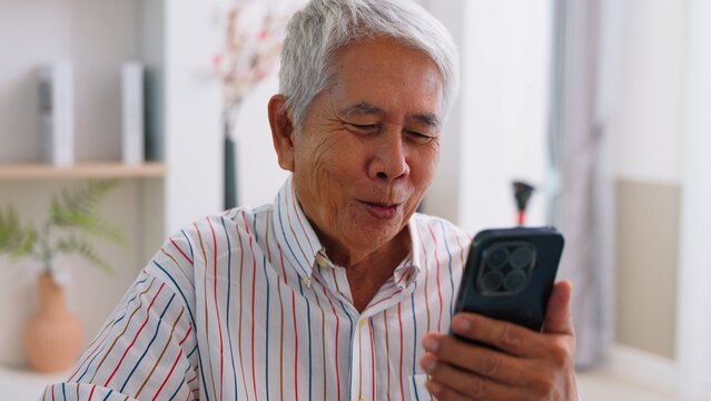 Happy senior Asian grandfather using smartphone for video call or watching online content at home. Cheerful elderly man enjoying modern digital technology lifestyle connection.