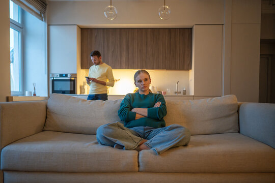 Emotional disconnection and ignorance in couple relationships, married partners ignoring each other, angry wife sitting on couch while uninterested manipulative husband wasting time on smartphone.