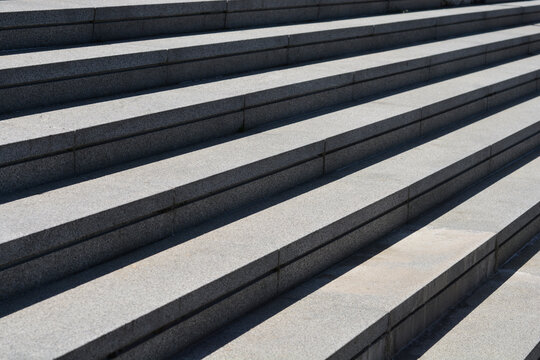 Diagonal view of wide gray granite outdoor stairs with repeating geometric lines and sharp linear shadows under bright sunlight