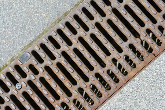 Diagonal view of a rusty metal storm drain grate channel set in a gray granite stone pavement structure under daylight