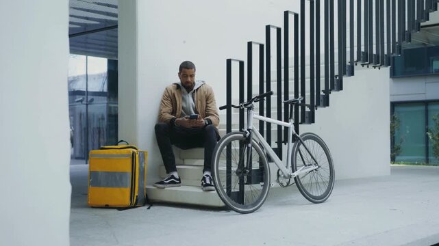 Tired courier sitting beside bicycle and yellow delivery bag near staircase