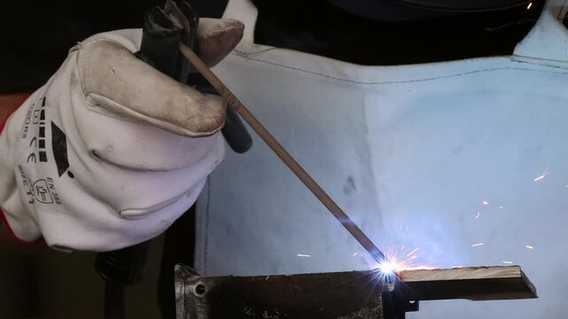 Professional welder in mask and protective apron welding metal workpiece with sparks