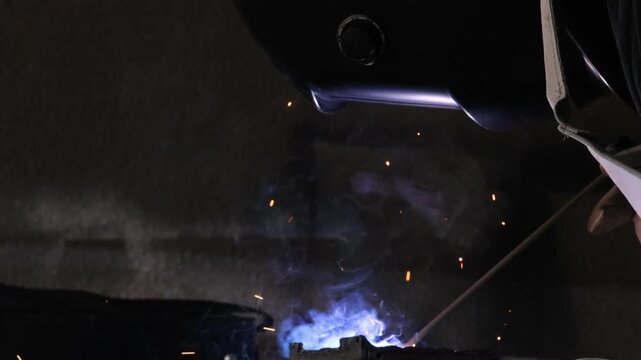 Close up of a welder hands in gloves holding electrode holder with flying sparks in dark workshop