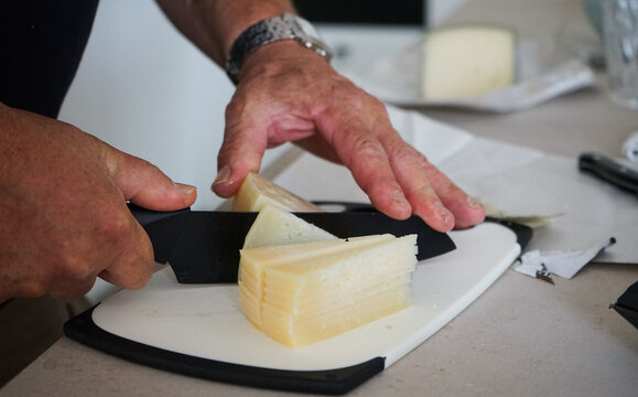 Primer plano de las manos de un hombre cortando queso curado con un cuchillo negro sobre una tabla.