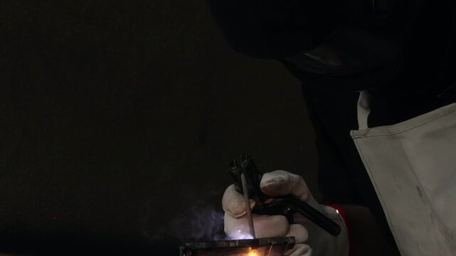 Close up of a welder hands in gloves holding electrode holder with flying sparks in dark workshop