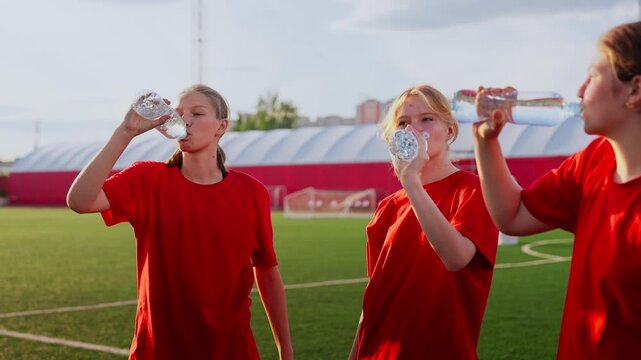 Womens soccer team hydrating and recovering during outdoor training session on green pitch under evening sunlight, wearing matching red jerseys and holding water bottles