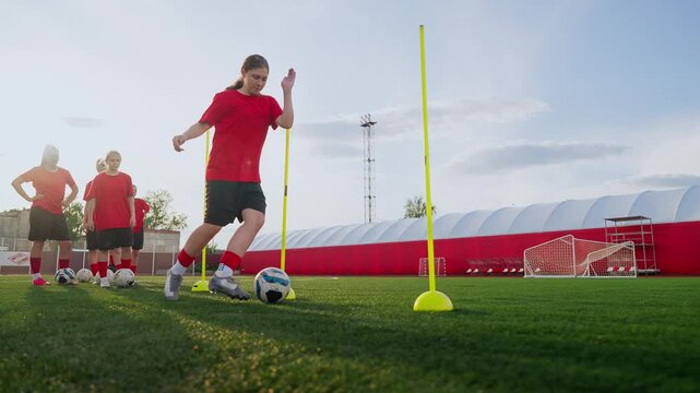 Focused women soccer players practicing dribbling and agility drills on green training field with cones, dummies and goals during organized outdoor practice session