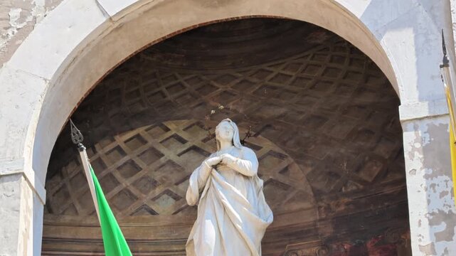 statue of Holy Mary standing gracefully on a crescent moon, positioned within an alcove featuring painted motifs and historic architecture of the clock tower of town hall in Modena, Italy