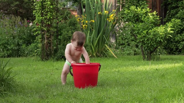 A toddler plays in the backyard. A red bucket is filled with toys and warm water. He takes turns taking out toys and examining them with a smile on his face. Italy