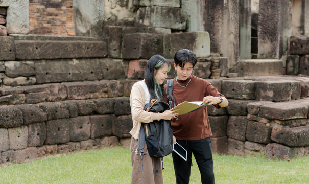 Happy Asian young man and woman tourist couple with backpack checking paper map and holding digital tablet while traveling together at historical ancient stone castle site.
